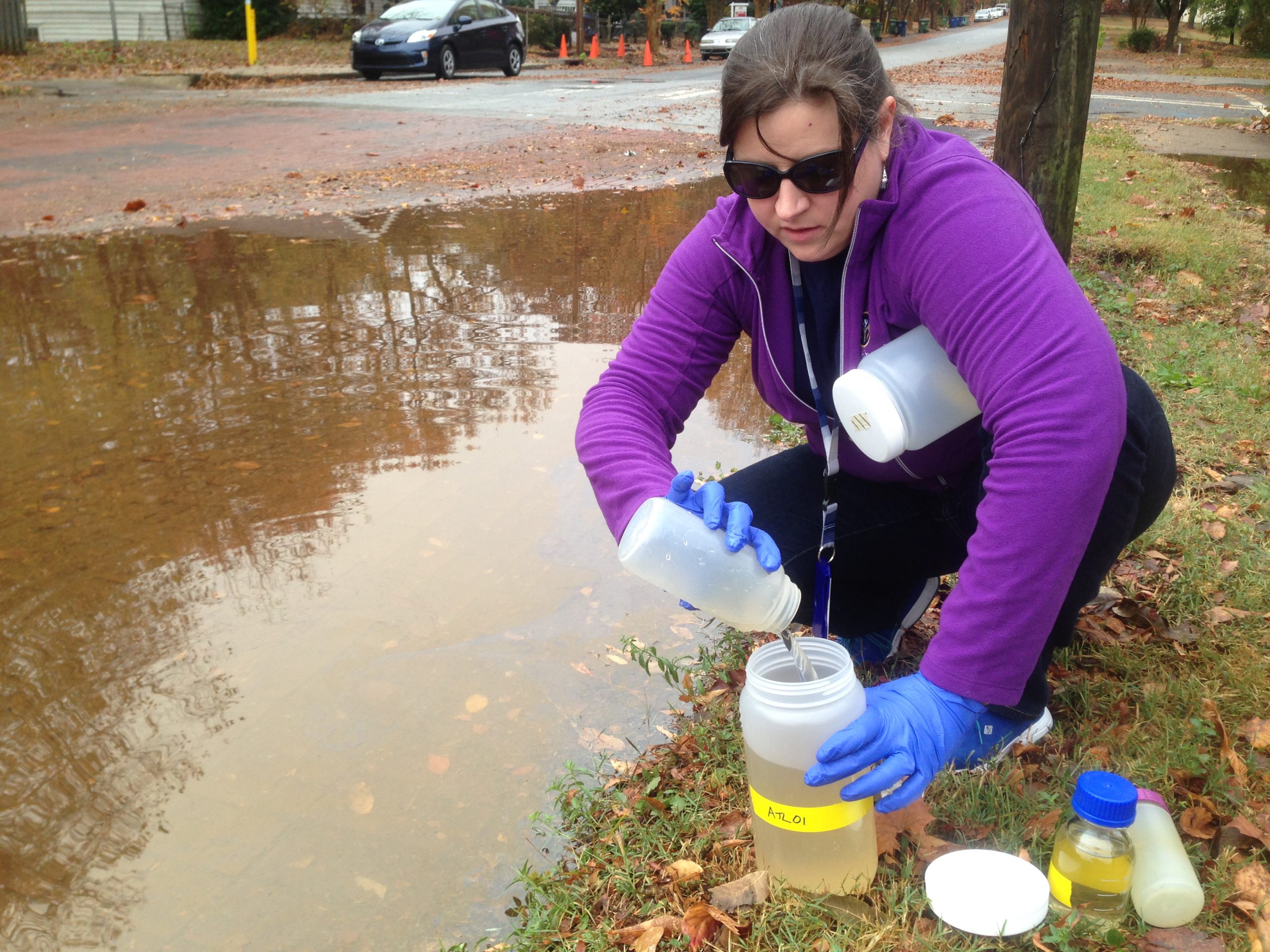 A photo of CDC microbiologist Amy Kirby kneeling next to a large puddle of floodwater pouring a sample into a large container.