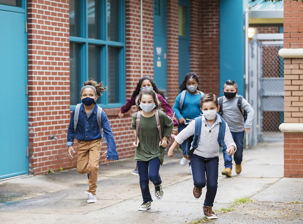 six children wearing masks and backpacks run in a schoolyard