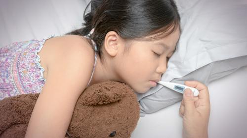 A young female child in bed with her eyes closed hugging a teddy bear while an adult's hand holds a thermometer in her mouth to check her temperature.