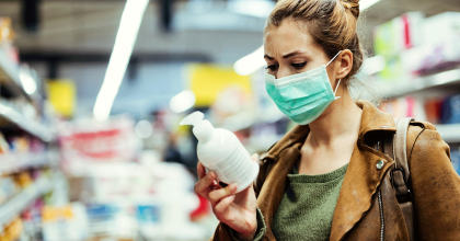 Young woman with protective mask reading label on hand sanitizer bottle while in the supermarket during pandemic.