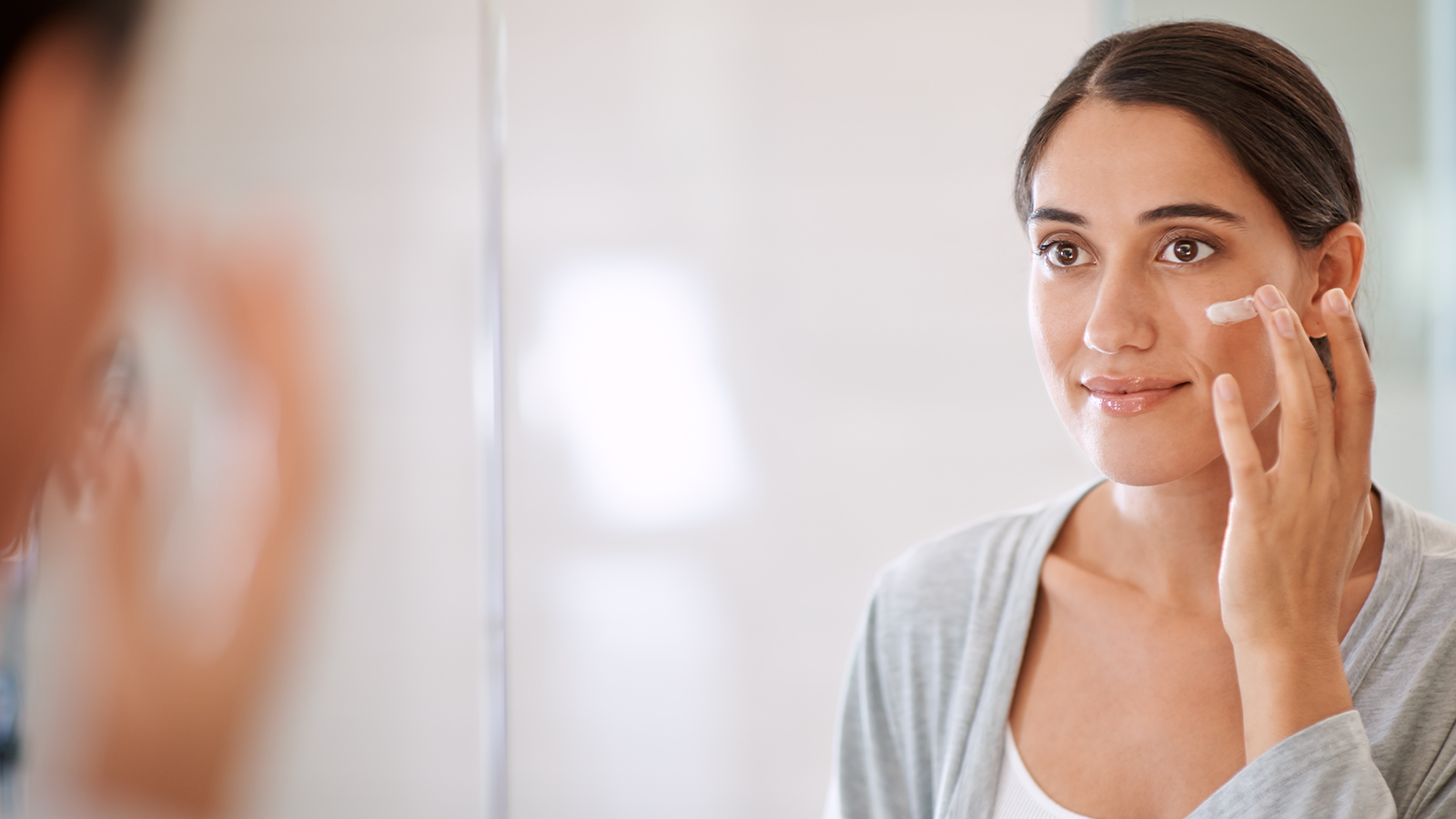 Woman applying face cream on her cheek bone while looking in a mirror