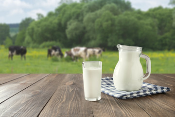 A table placed outside with a pitcher and glass on it, both full of milk. In the distance are rolling hills with cows.