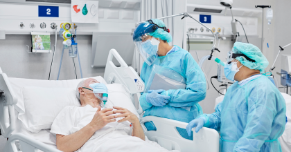 Man lays in a hospital bed connected to ventilator looking up at nurses in blue scrubs and personal protective equipment 