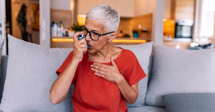 Senior short gray-hair woman wearing eyeglasses using asthma inhaler while sitting on sofa ta home during the day.