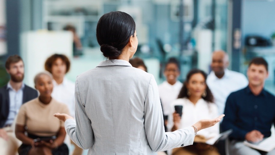 Photo of a woman standing at the front of the room, and she is speaking to a group of people. 