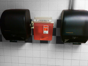 Sharps disposal container mounted in a restroom between two towel dispensers