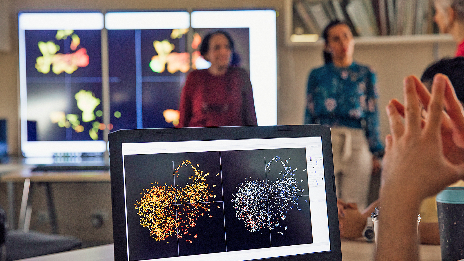 A close up of a laptop screen can be seen depicting a scientific image. A similar image is repeated on large screens towards the back of the photograph. A pair of hands can be seen in the foreground and there are some students in the background who are out of focus.