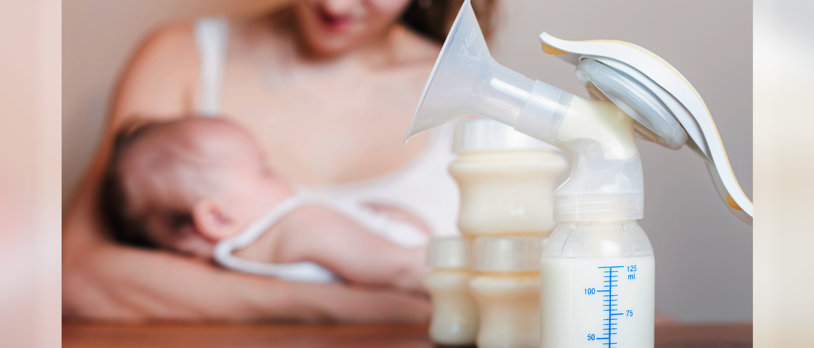 a breast pump with bottles of breast milk on a table in the foreground, and a young woman cradling a baby in her arms in the background