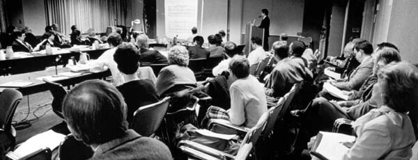 An advisory committee meeting: A man giving a presentation at a lecturn, committee members sitting around a conference table, and observers sitting in rows of chairs