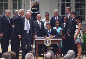 color image President Obama sitting at desk surrounded by people signing document