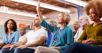 A woman is sitting in an audience composed of several people and she is raising her hand.