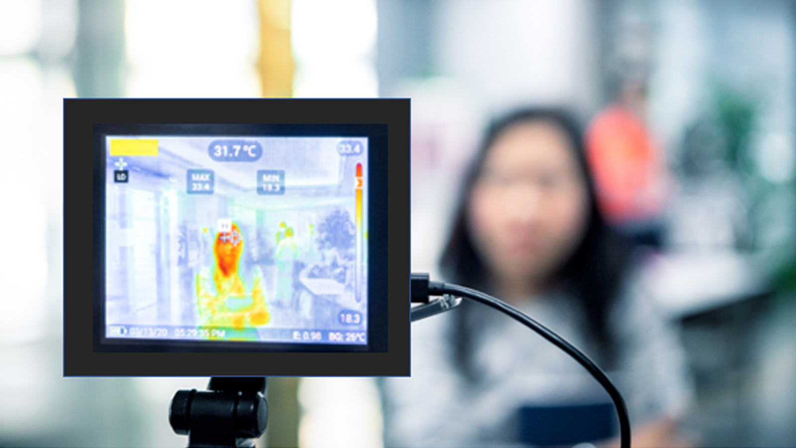 Figure one: A picture of an infrared thermal camera pointed at a woman standing by herself in a public space. The camera displays her thermal image on the camera screen. Her face is shown on the screen in a reddish orange color indicating her skin has a higher surface temperature than her clothing displayed as yellow and the distant background displayed as gray. The temperature displayed on the screen as 31.7o Celsius. 
