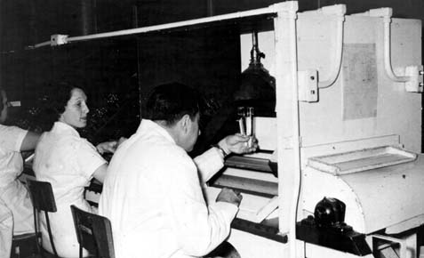Man in a white lab coat seated at a piece of manufacturing equipment examining a vial with a woman looking on