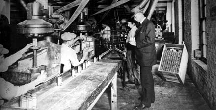 Two workers in a manufacturing plant packing bottles of product with a man in a suit and hat looking on