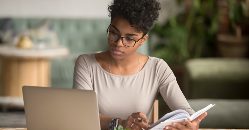 Young woman reading, holding a book and looking at a laptop computer