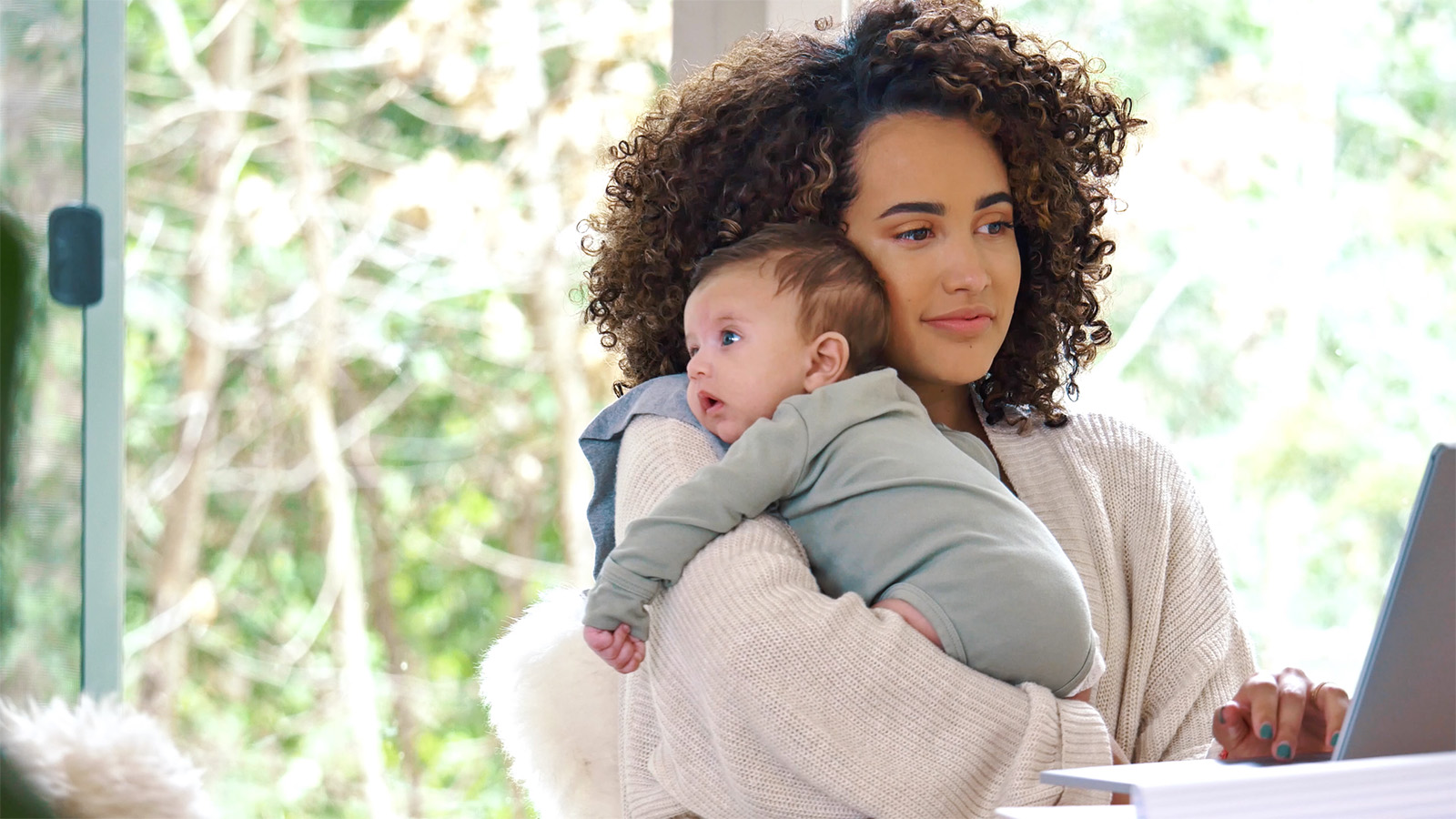 Image of young mother working on a laptop while carrying an infant