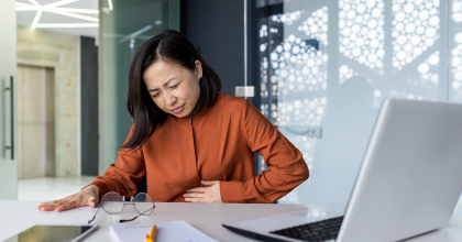 Sick Asian business woman at workplace holding hand on stomach from acute pain 