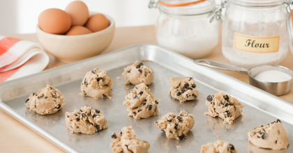 Photo of raw cookie dough on baking tray on kitchen counter with eggs, sugar, and flour in the background.