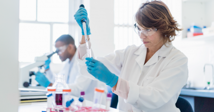 Female lab technician running tests in lab with male coworker in background looking through microscope.