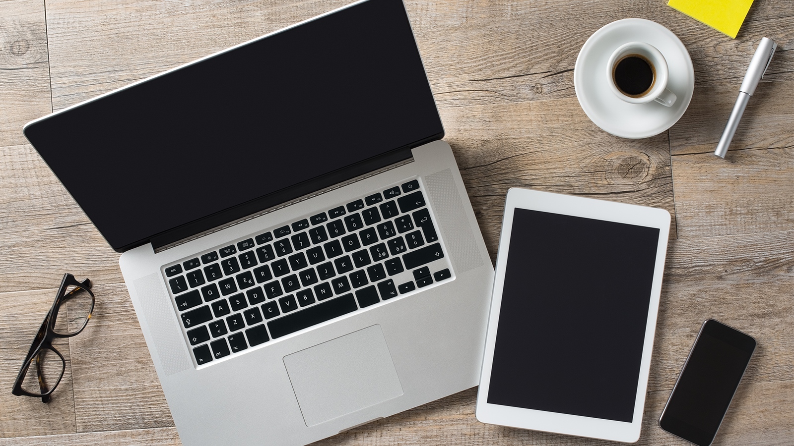 Open Laptop with glasses to the left and coffee cup, pen, tablet and phone on the right