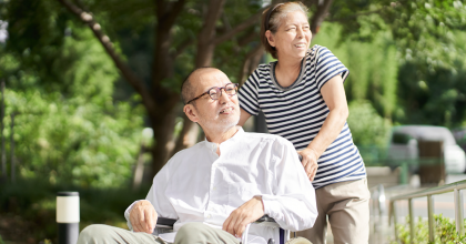 A man wearing glasses and a white shirt is in a wheelchair being pushed by a woman caretaker.