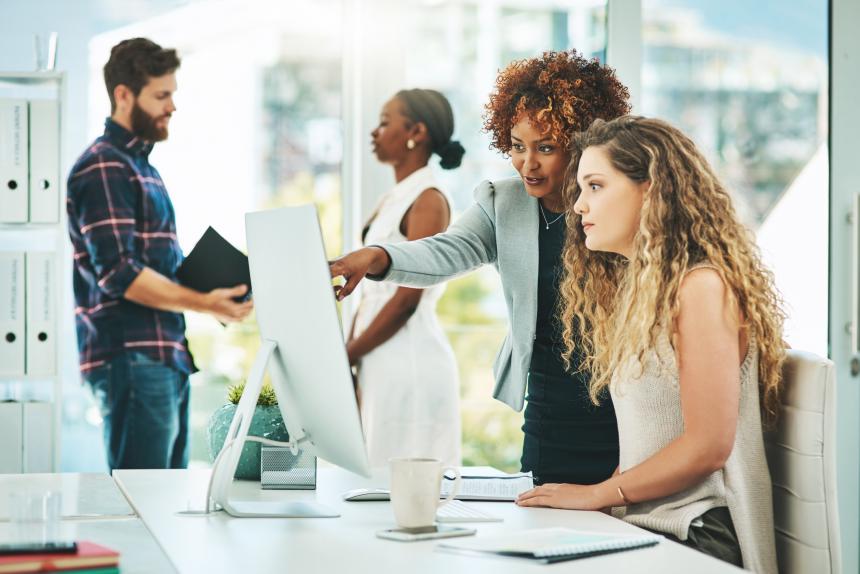 Image of ladies looking a computer screen while working together on a project. The image of man and women in the background talking 