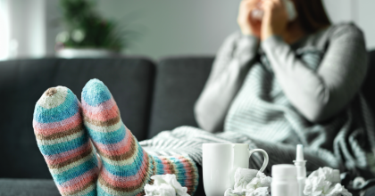 Woman sitting on couch blowing her nose, surrounded by used tissues, cup of tea, nasal spray, and bottle of medicine.