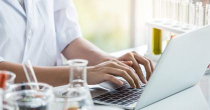 Person in a lab coat typing on a laptop with beakers in the foreground.