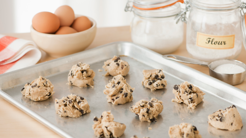 Photo of raw cookie dough on baking tray on kitchen counter with eggs, sugar, and flour in the background.